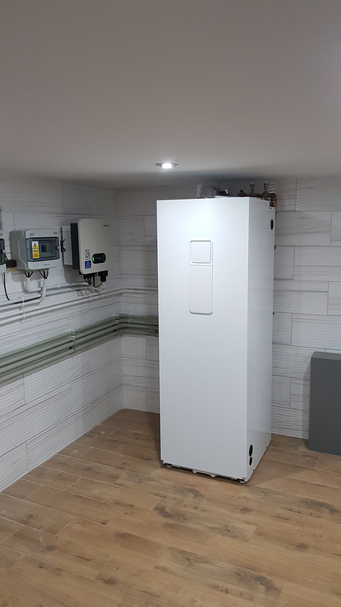 Biały, prostopadłościenny zasobnik ciepłej wody użytkowej w rogu pomieszczenia z panelami fotowoltaicznymi i rozdzielnicą elektryczną na ścianie, podświetlony punktowym światłem.
