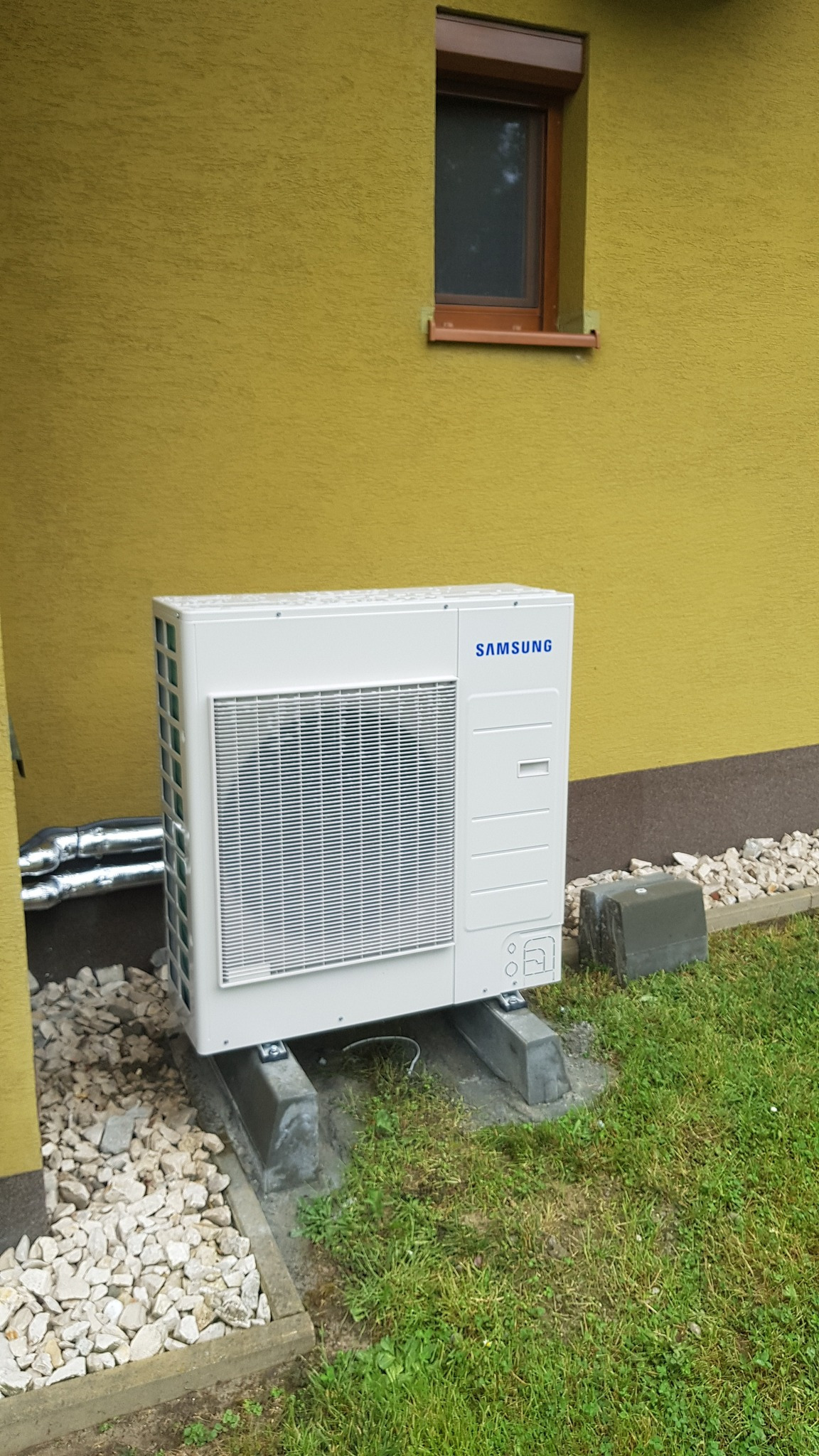 Biała, zewnętrzna jednostka klimatyzacji Samsung zamontowana na betonowych podstawach przy żółtej ścianie budynku z małym oknem i brązową ramą, otoczona białym żwirem i zieloną trawą.