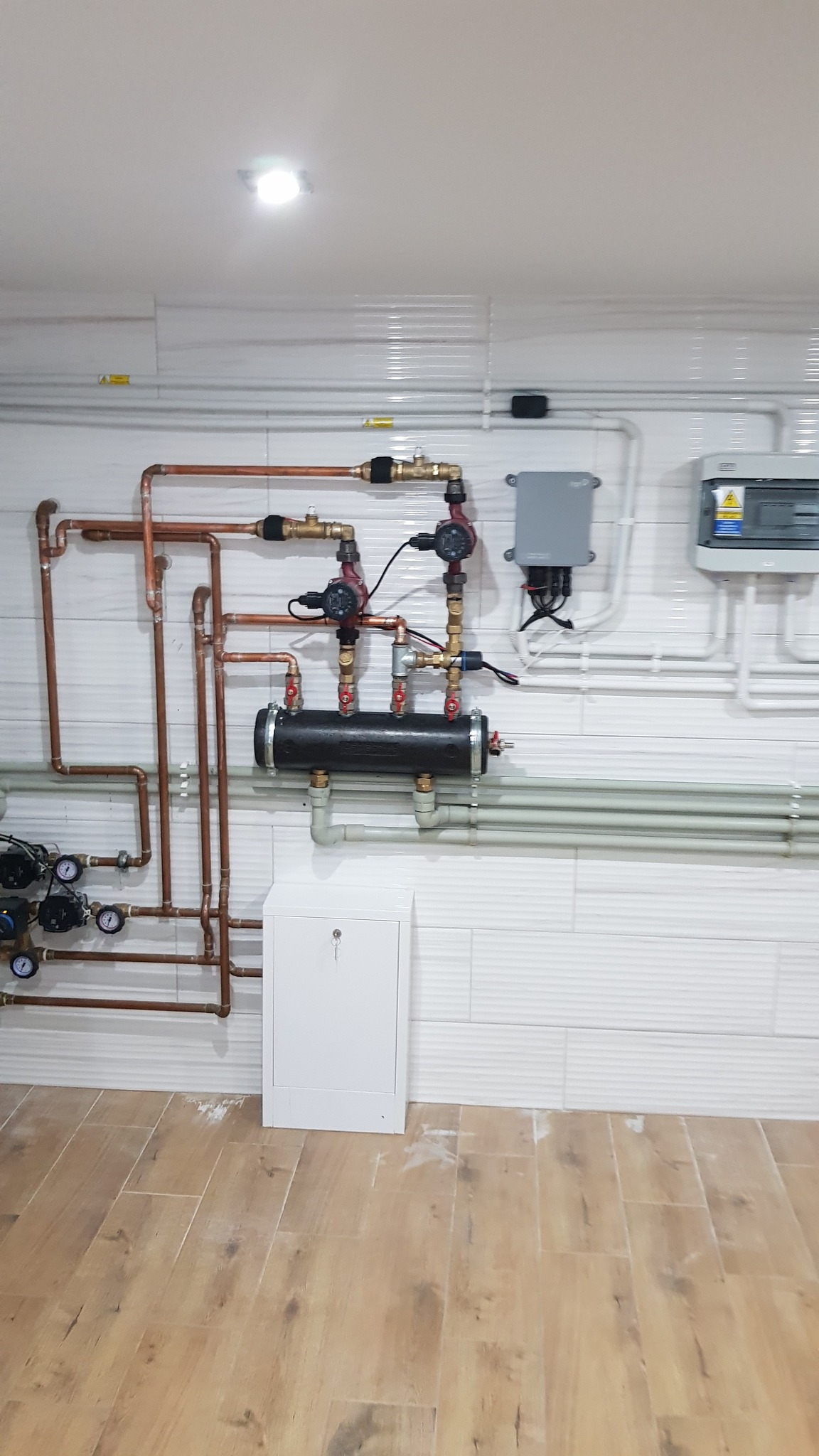 Rozbudowana instalacja centralnego ogrzewania z licznymi rurami miedzianymi i elementami sterującymi, zamontowana na ścianie wyłożonej białymi płytkami imitującymi cegłę, z widoczną szafką zamykaną...