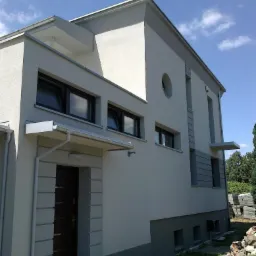 Elewacja domu w odcieniach szarości z nowoczesnymi akcentami architektonicznymi, okrągłym oknem i stertą bloczków obok. Ujęcie z dołu, błękitne niebo w tle.