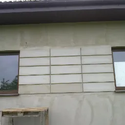 Fragment elewacji budynku po nałożeniu warstwy tynku strukturalnego i dekoracyjnych paneli betonowych. Widoczne okna i paleta budowlana u dołu.