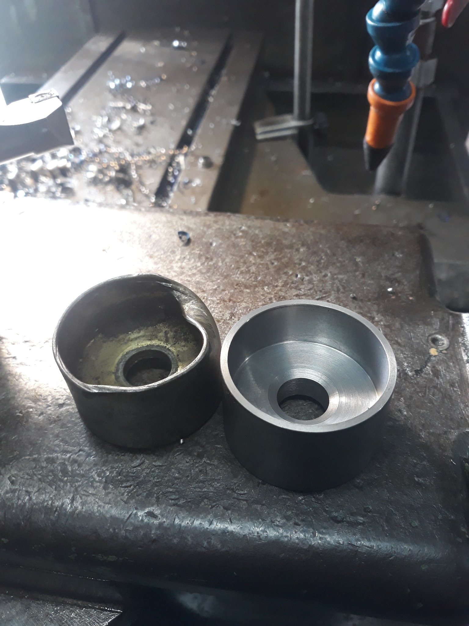 Dwie metalowe tuleje, jedna surowa z widocznymi śladami obróbki i niedoskonałościami, druga precyzyjnie obrobiona z gładką powierzchnią, leżą na brudnym stole warsztatowym z wiórami i resztkami po...