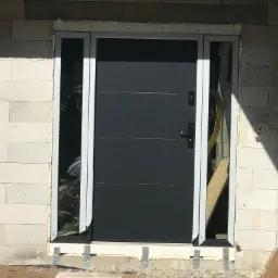 Montaż czarnych drzwi zewnętrznych z bocznymi okienkami w surowym stanie budynku w Jeleniej Górze. Widoczne elementy montażowe i pianka uszczelniająca.