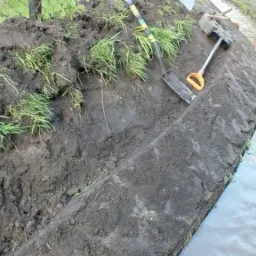 Uformowany brzeg cieku wodnego z użyciem ziemi, widoczne ślady pracy łopatą i fragmenty darni, w tle ogrodzenie z siatki.