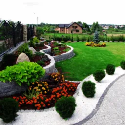 Urozmaicony ogród z falistymi rabatami, kamiennymi murkami, bujną zielenią, kolorowymi kwiatami i równo przystrzyżonym trawnikiem, widok z góry w kierunku domu i oddalonego miasteczka.