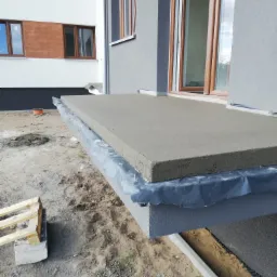 Szara wylewka na balkonie w trakcie prac wykończeniowych, zabezpieczona czarną folią, widok z boku, w tle fragment elewacji budynku.