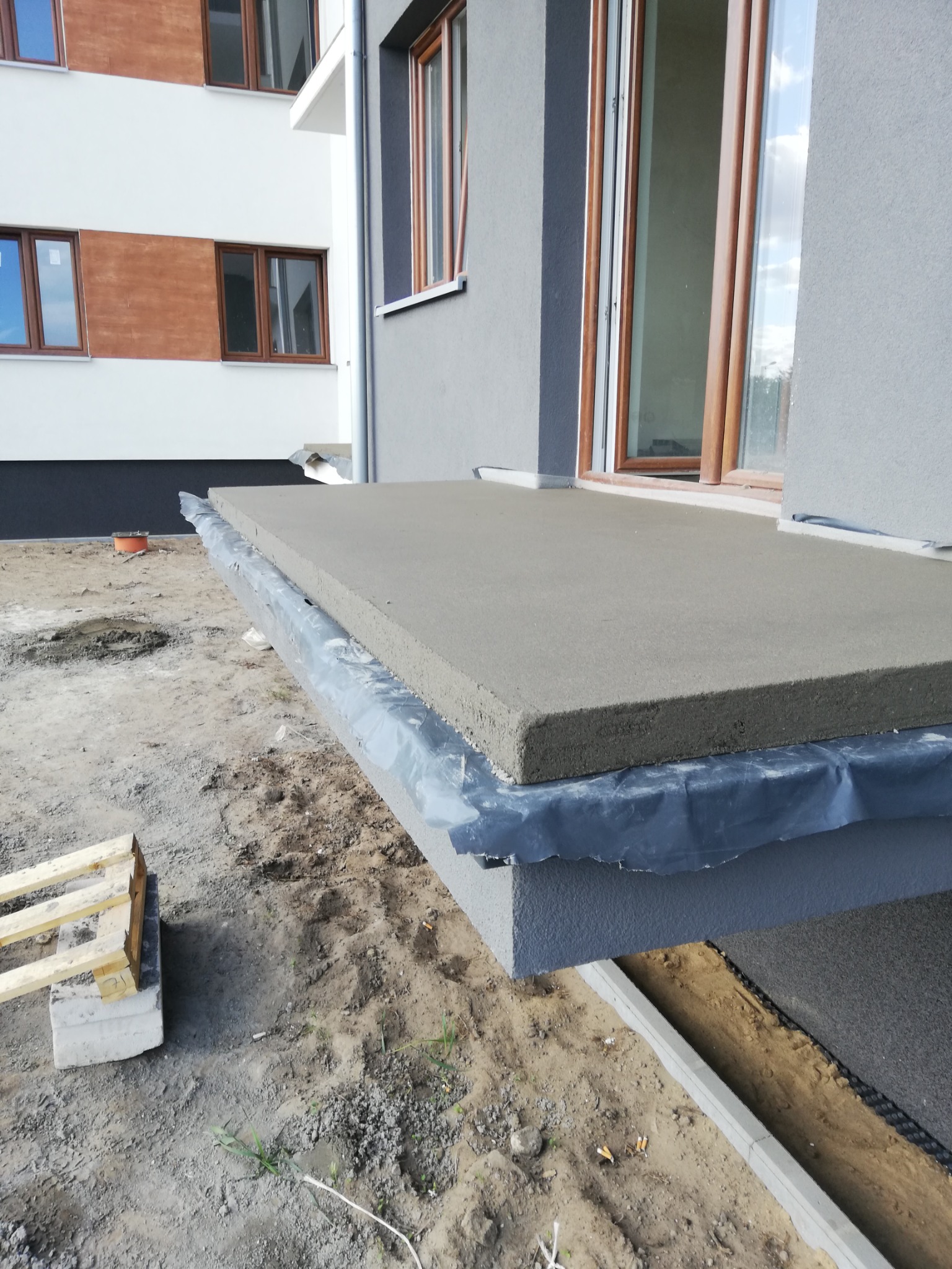 Szara wylewka na balkonie w trakcie prac wykończeniowych, zabezpieczona czarną folią, widok z boku, w tle fragment elewacji budynku.