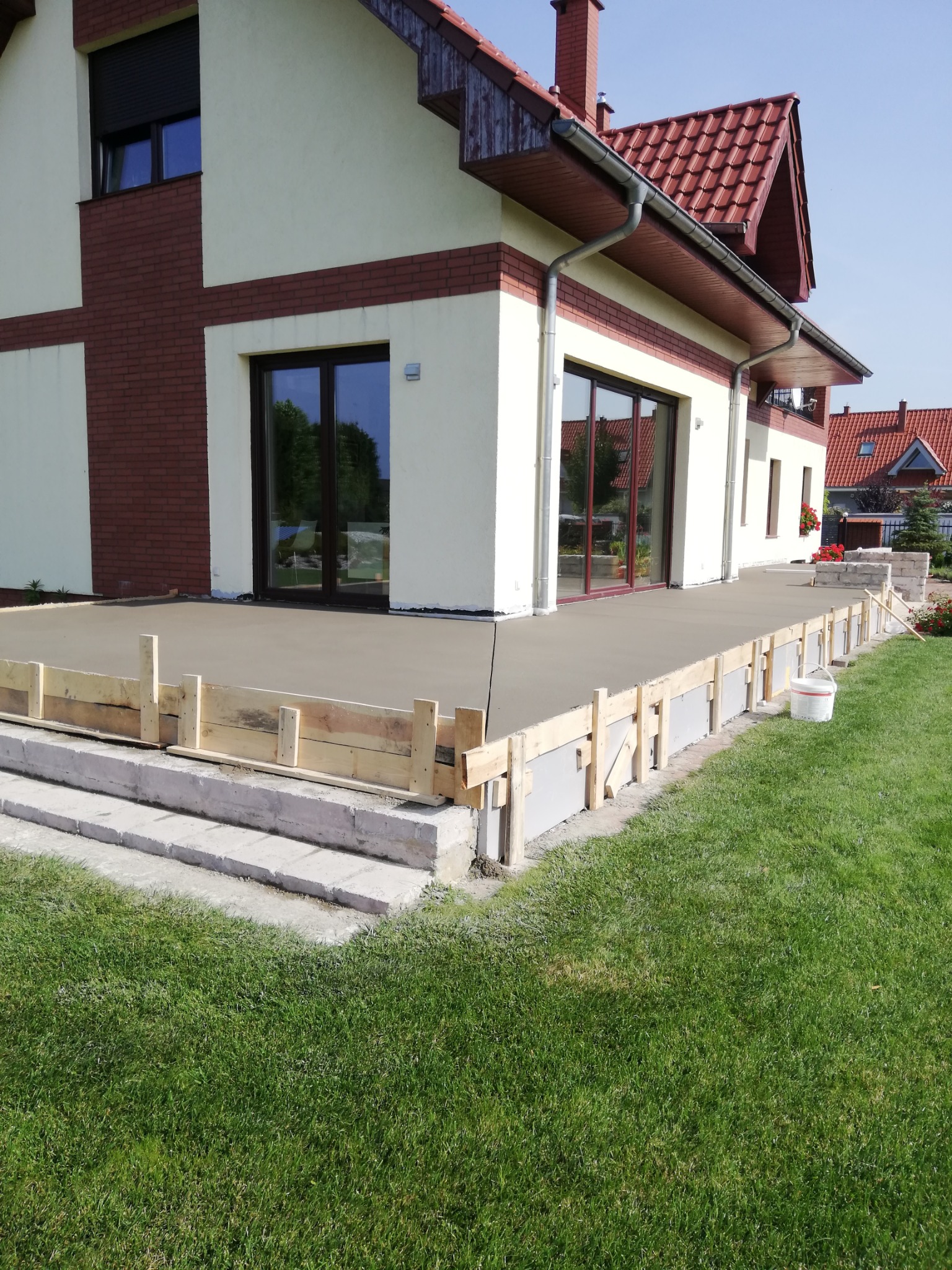 Wylewka jastrychowa na tarasie domu jednorodzinnego w trakcie realizacji, widoczne drewniane szalunki, jasny kolor wylewki, trawnik, biały budynek z elementami cegły