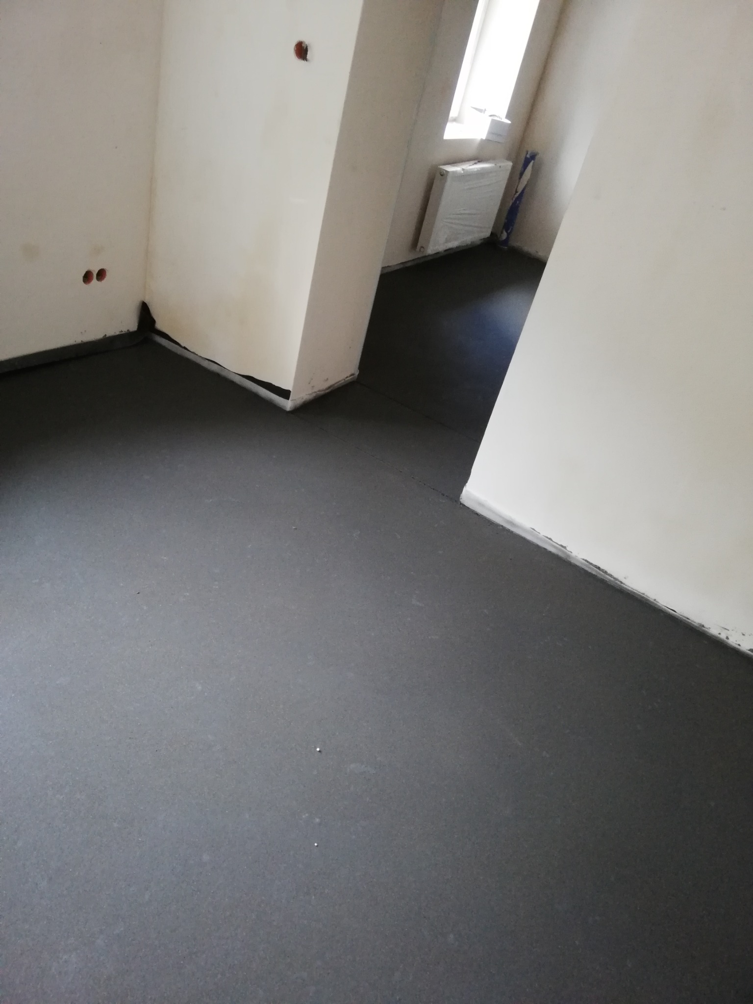 Świeżo wylana, ciemnoszara posadzka w surowym wnętrzu, widoczne ślady po listwach przypodłogowych i otwory po gniazdkach elektrycznych.