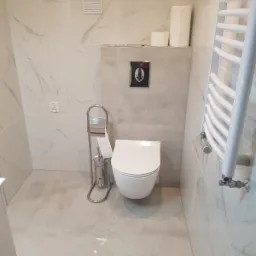 Nowoczesna toaleta wisząca z chromowanym przyciskiem spłukującym, obok biały grzejnik drabinkowy i stojak na papier toaletowy z szczotką, marmurowe płytki na ścianach i podłodze, wentylator...