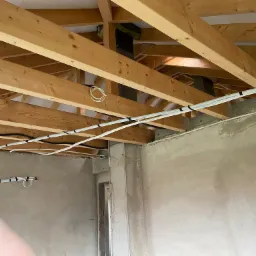 Surowy stan wnętrza domu w trakcie instalacji przewodów wentylacyjnych pod drewnianą konstrukcją dachu, z widocznym kominem i otynkowanymi ścianami.