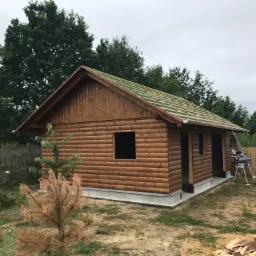 Drewniany domek w trakcie budowy z częściowo ułożonym dachem, widoczne krokwie i narzędzia na zewnątrz.