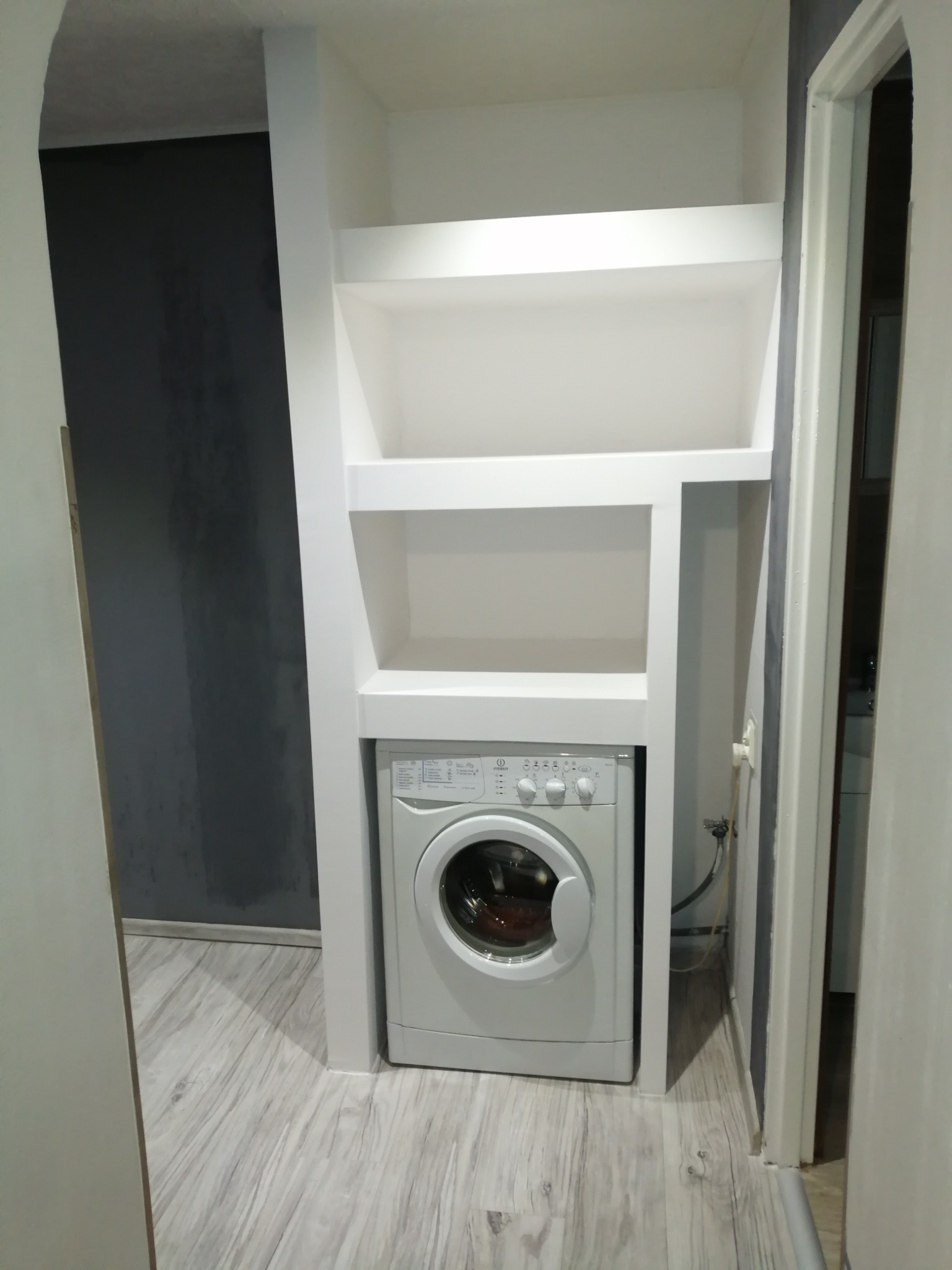 Białe, zabudowane półki z płyty gipsowo-kartonowej nad pralką w łazience z szarą ścianą i panelami podłogowymi.