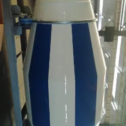 Bęben betoniarki pomalowany w biało-niebieskie pasy, zawieszony na konstrukcji w hali produkcyjnej, widoczny fragment kabiny samochodu