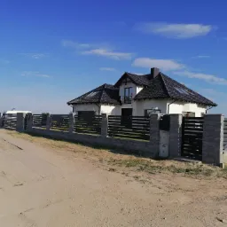 Nowoczesne ogrodzenie panelowe z poziomych, grafitowych przęseł pomiędzy słupkami z szarej kostki, chroniące posesję z białym domem z czarnym dachem w tle, widoczne skrzynki elektryczne przy furtce.