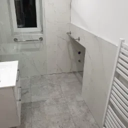 Nowoczesna łazienka z szarymi płytkami podłogowymi i białymi płytkami ściennymi imitującymi marmur, wyposażona w białą szafkę umywalkową, grzejnik drabinkowy oraz uchwyty dla niepełnosprawnych przy...