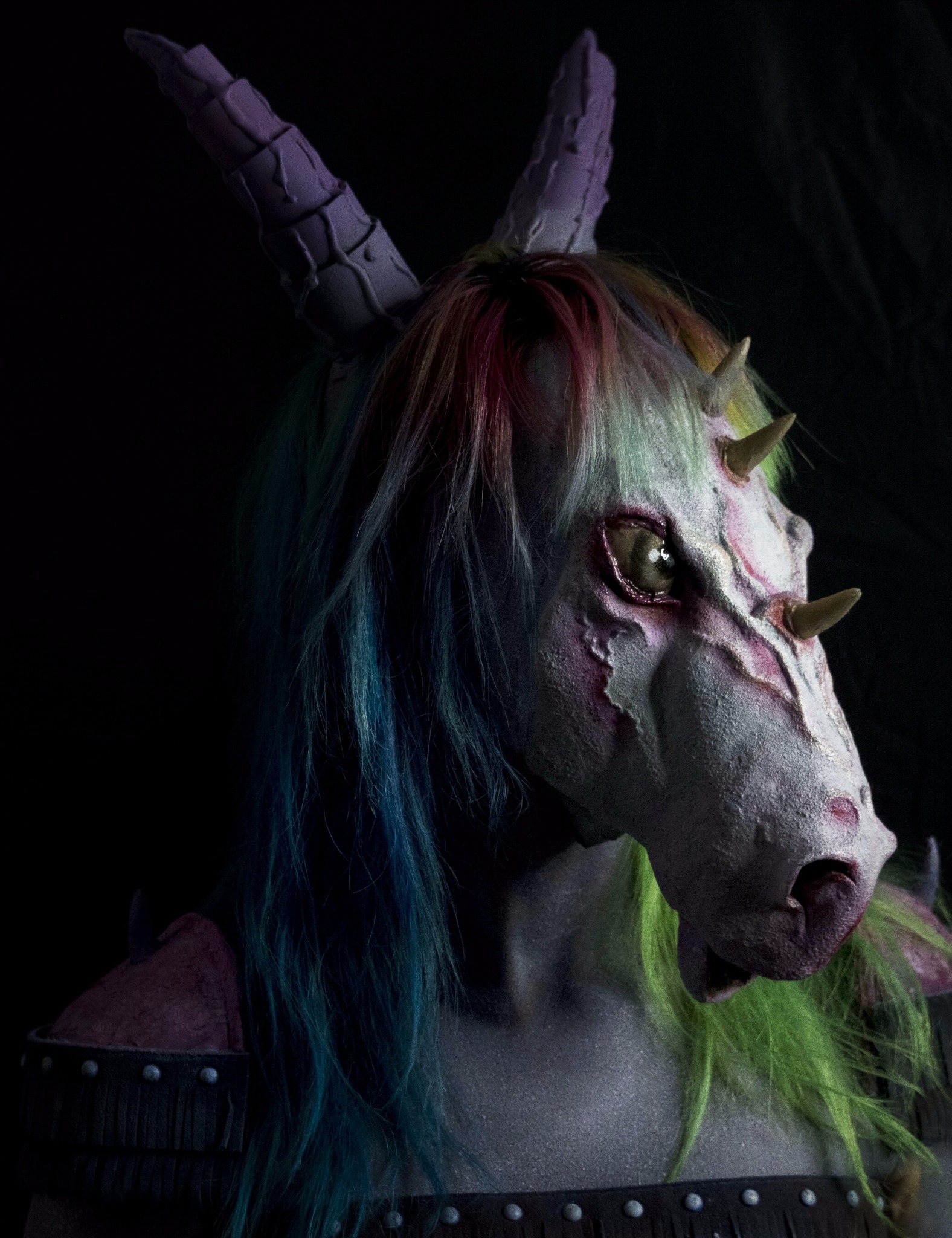 Profilowe ujęcie osoby w fantazyjnej masce jednorożca z tęczową grzywą i fioletowymi rogami, w kostiumie z elementami zbroi, na ciemnym tle.