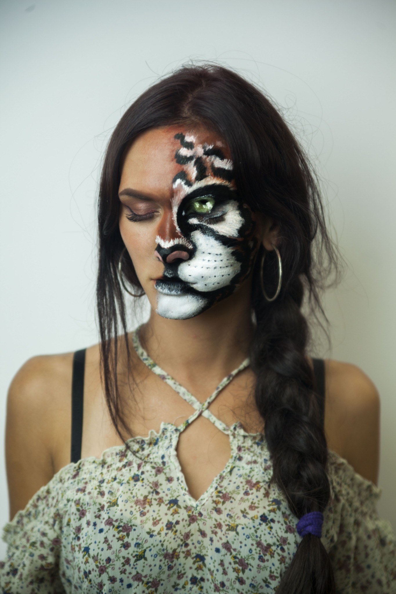 Kobieta z artystycznym makijażem twarzy, przedstawiającym motyw tygrysa na połowie twarzy, z zielonym okiem i czarnymi paskami, spleciony warkocz z fioletową gumką