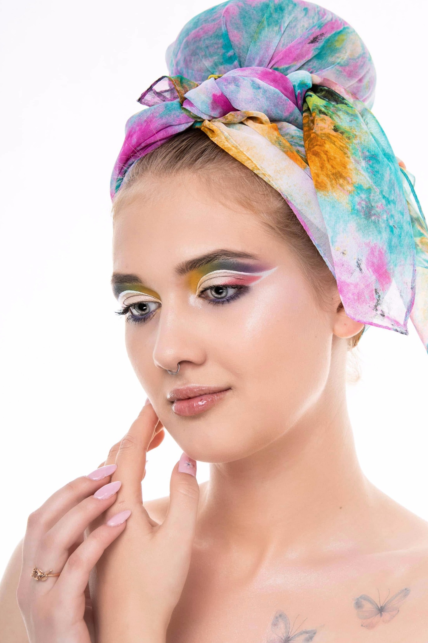 Portret kobiety z awangardowym makijażem, kolorową chustą na głowie, piercingiem w nosie i tatuażami motyli na dekolcie, pozującej na białym tle.