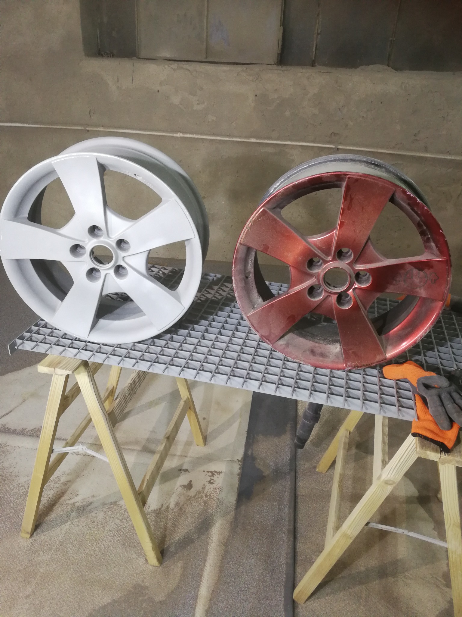 Dwa aluminiowe felgi samochodowe na metalowej kratce: jedna po piaskowaniu i malowaniu na biało, druga w kolorze czerwonym przed obróbką, obok leżą rękawice robocze.