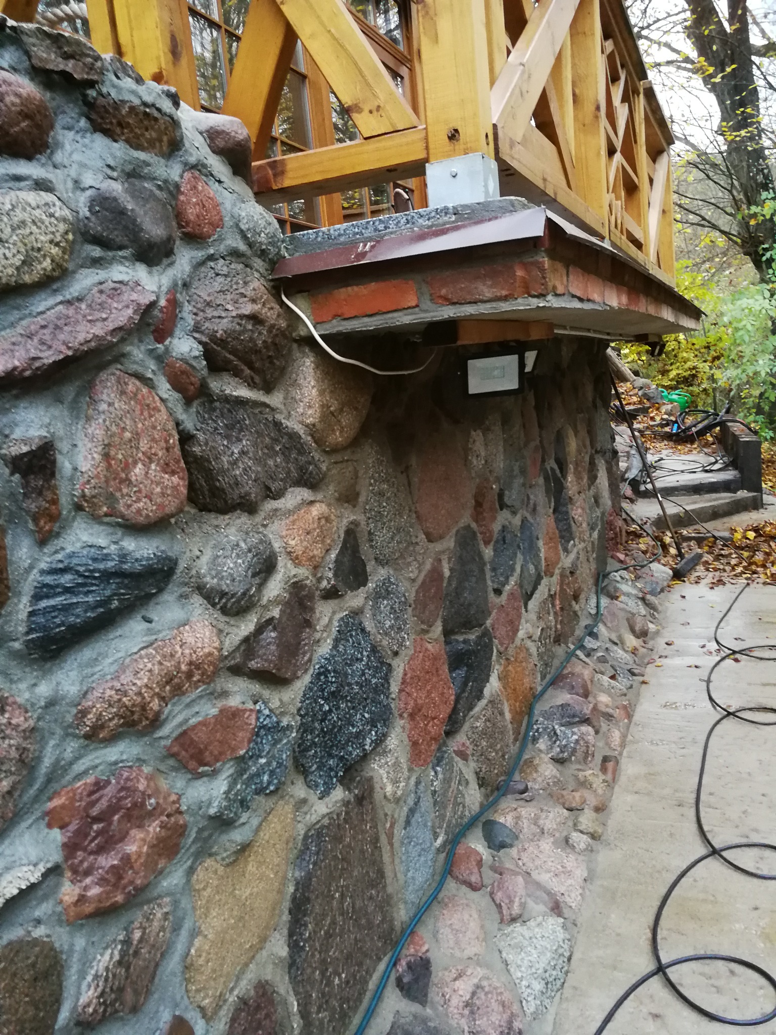 Kamienny mur z drewnianą konstrukcją u góry, widoczne przewody elektryczne i reflektor na ścianie, otoczenie z liśćmi i schodami.