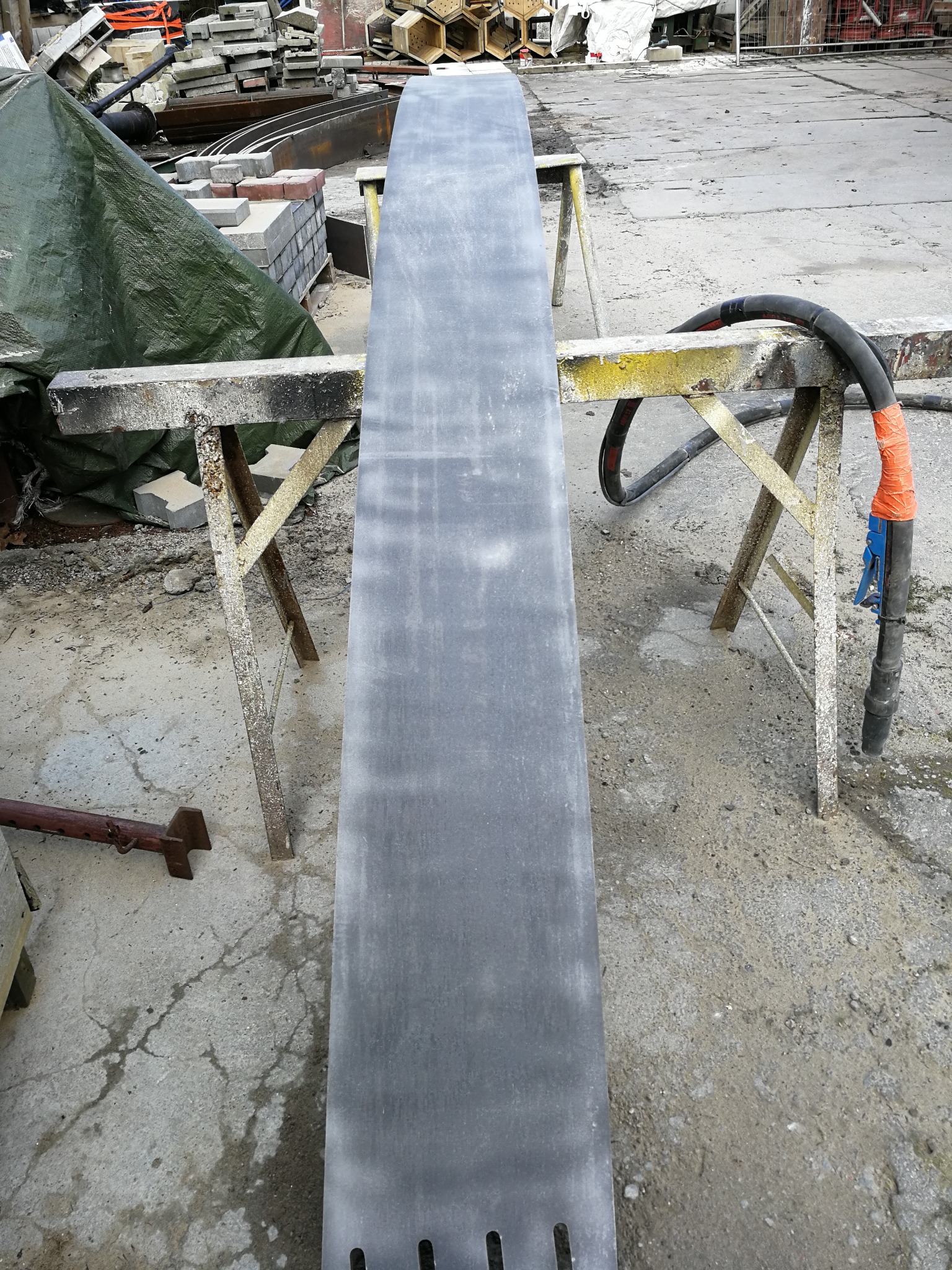 Długa, szara płyta metalowa na dwóch metalowych kozłach, z widocznymi śladami obróbki powierzchni, obok węża z dyszą do czyszczenia pod ciśnieniem.