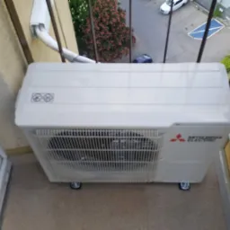 Biała jednostka zewnętrzna klimatyzatora Mitsubishi Electric na balkonie, widok z góry, z widocznymi elementami konstrukcyjnymi balkonu i fragmentem zieleni na zewnątrz.