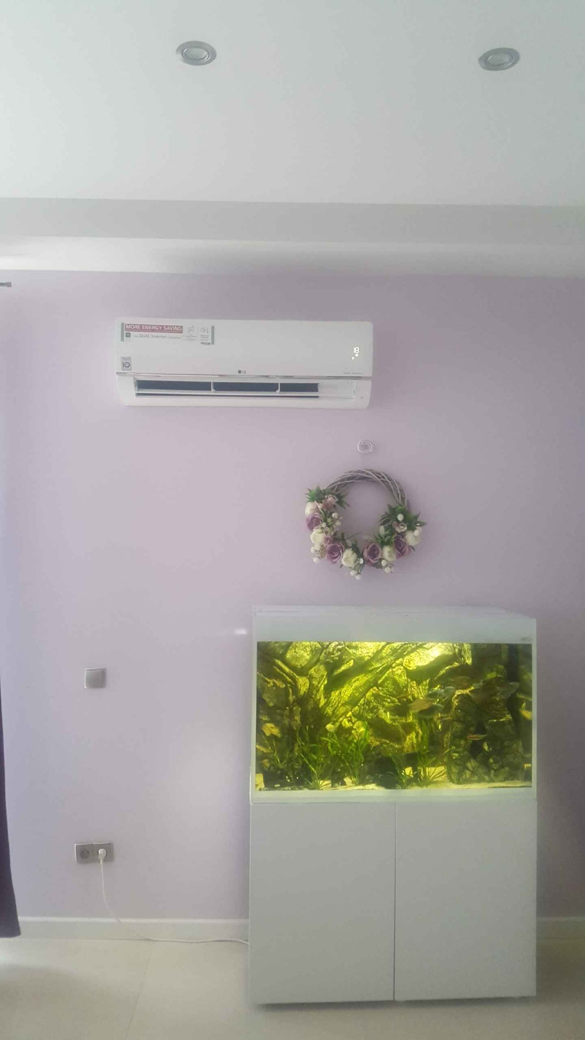Biała jednostka klimatyzacyjna LG zamontowana na liliowej ścianie nad akwarium z roślinami w Warszawie, ozdobiona wiankiem z kwiatów.