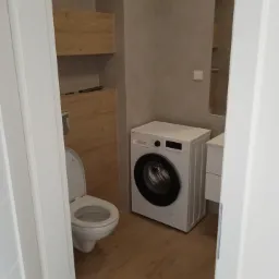 Wykończona łazienka w odcieniach beżu i bieli, z wiszącą toaletą i pralką automatyczną, widok zza uchylonych drzwi.