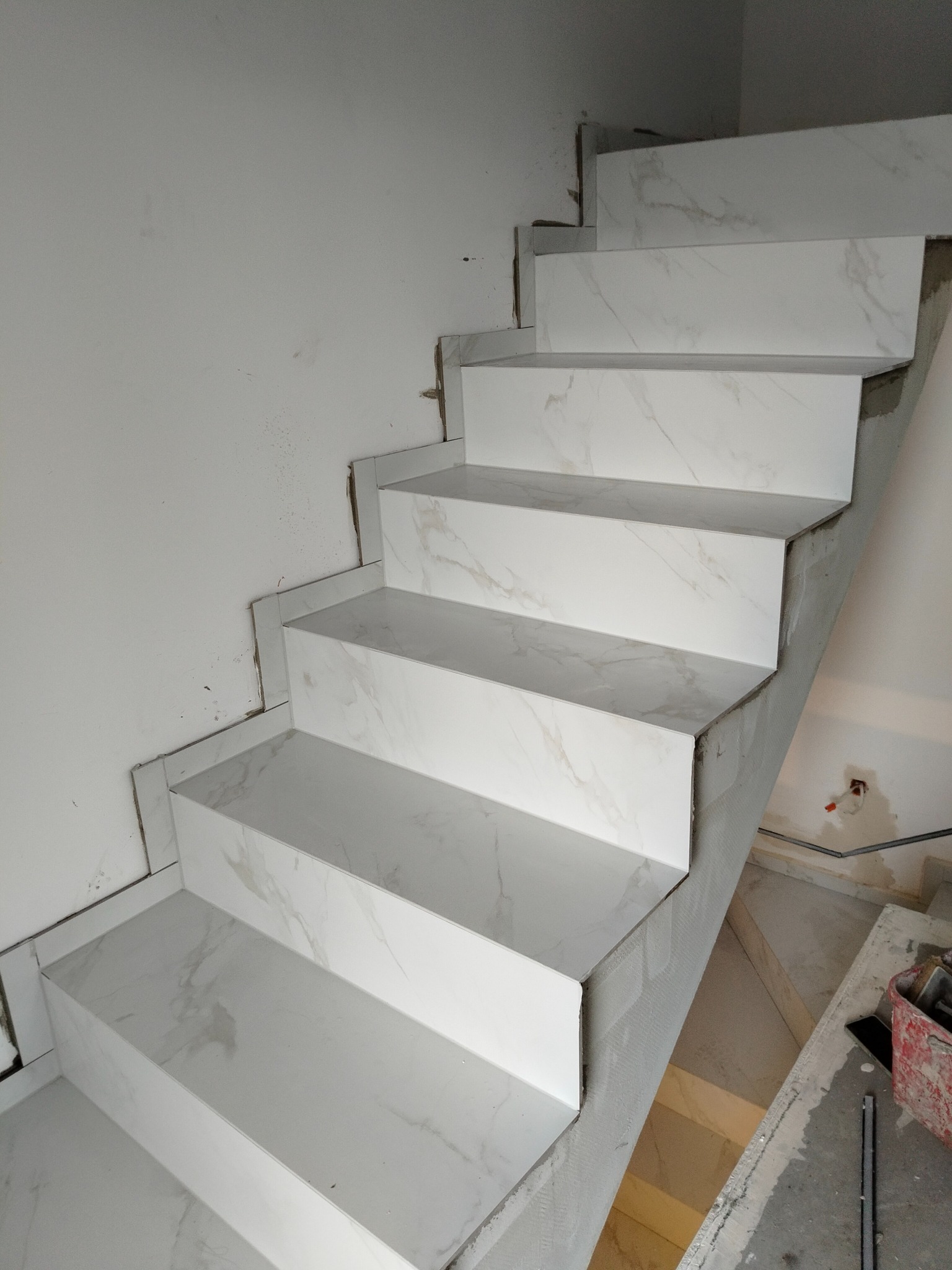 Schody wyłożone białymi płytkami imitującymi marmur, widoczne niedokończone prace wykończeniowe na ścianie i podłodze. Ujęcie z dołu, podkreślające wzór płytek.