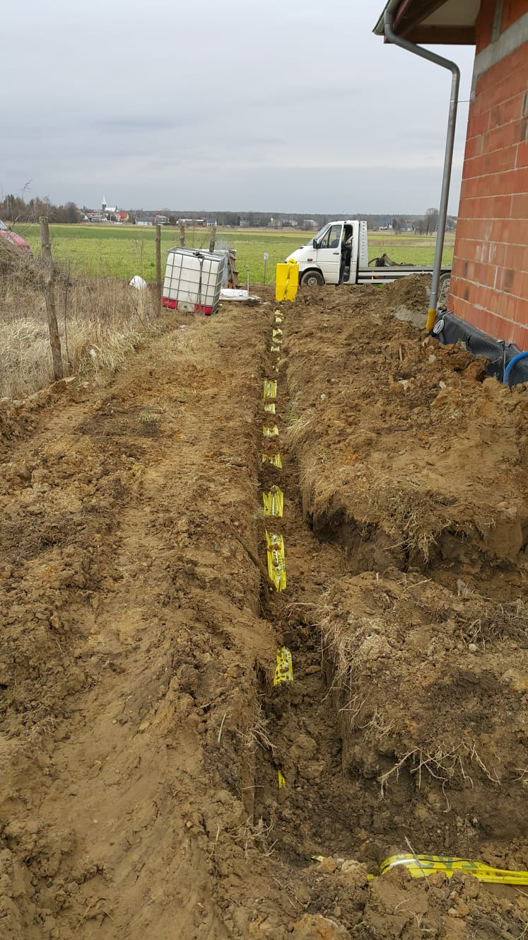 Wykop pod instalację sanitarną z widoczną taśmą ostrzegawczą w Tarnowskich Górach, w tle budynek w stanie surowym i biały samochód dostawczy.