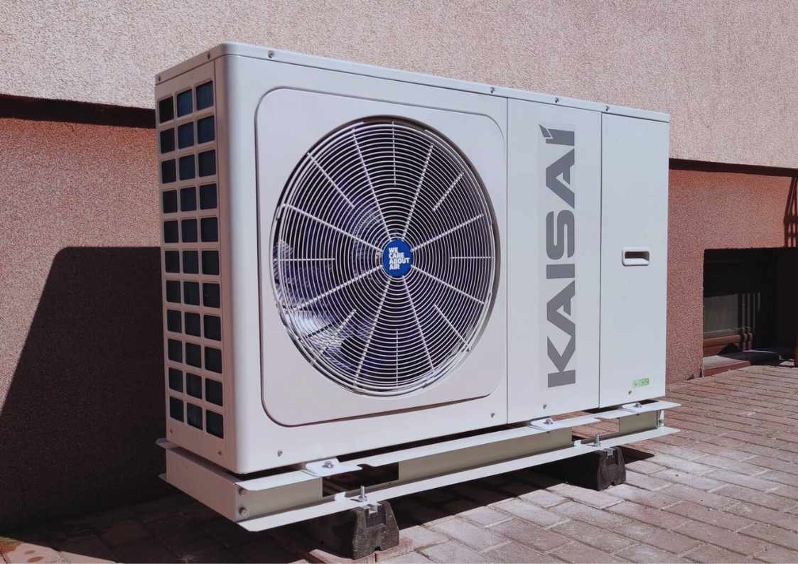 Biała pompa ciepła Kaisai zamontowana na zewnątrz budynku, widoczna z boku na tle ceglanej ściany i kostki brukowej. Wentylator z niebieskim logo w centrum.