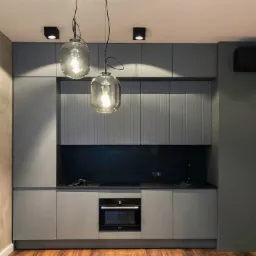 Nowoczesna szara kuchnia z drewnianą podłogą, oświetlona dwoma wiszącymi lampami o szklanych kloszach i punktowymi reflektorami sufitowymi, z czarnym panelem nad blatem i wbudowanym piekarnikiem.