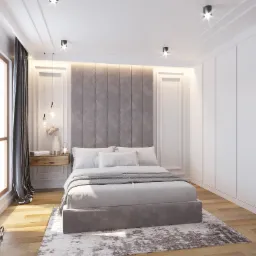Nowoczesna sypialnia w apartamencie: łóżko z szarym wezgłowiem, drewniana szafka nocna z lampkami i kwiatami, zasłony zaciemniające okno, dywan w odcieniach szarości i bieli, oraz biała zabudowa...