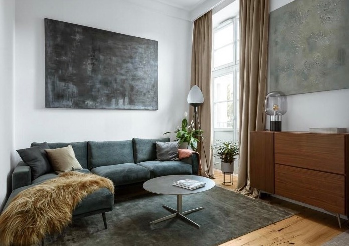 Nowoczesny salon z szarozieloną sofą, okrągłym stolikiem kawowym na metalowej nodze i dużym abstrakcyjnym obrazem w odcieniach szarości na ścianie. Na podłodze leży ciemnoszary dywan, a obok okna...