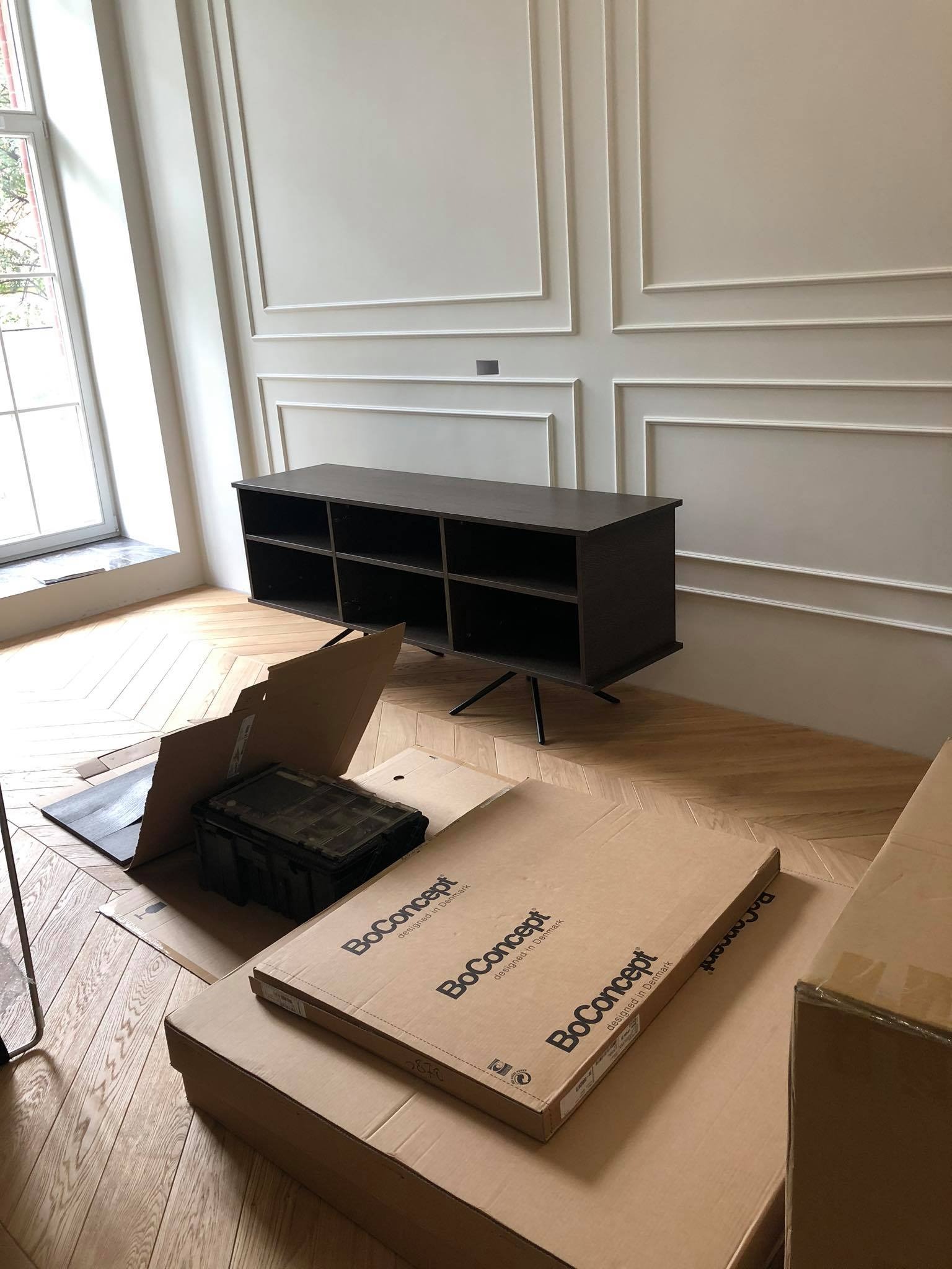 Montaż brązowego regału BoConcept w apartamencie z parkietem w jodełkę i sztukaterią na ścianach, widoczne kartony i narzędzia.