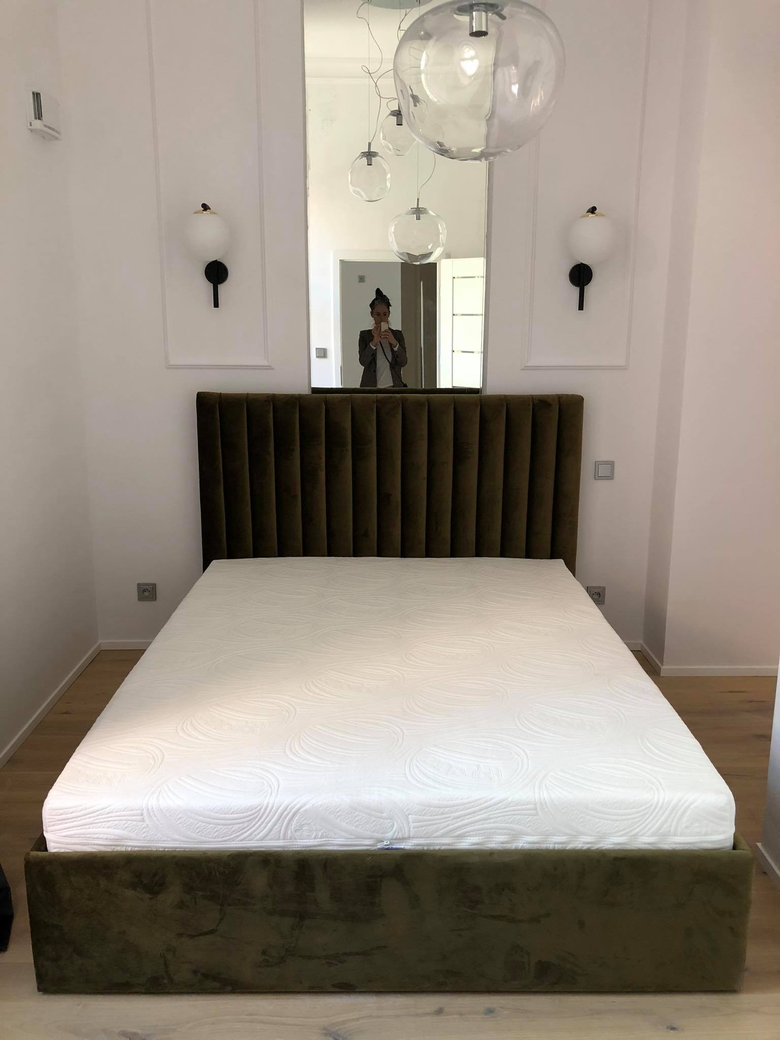 Zielone łóżko z welurowym zagłówkiem w minimalistycznej sypialni z białymi ścianami, oświetleniem kulistym i lustrem odbijającym postać robiącą zdjęcie.