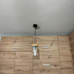 Szklana lampa wisząca z żarówką filamentową, zamontowana na tle drewnianej zabudowy meblowej.