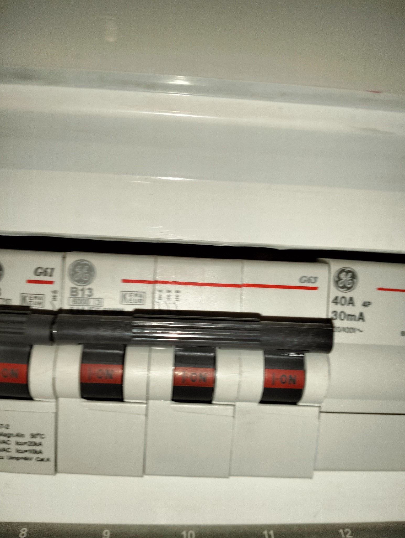 Zbliżenie na tablicę rozdzielczą z wyłącznikami nadprądowymi marki GE, widoczne oznaczenia 40A i 30mA, przełączniki w pozycji ON, numery obwodów 8-12.