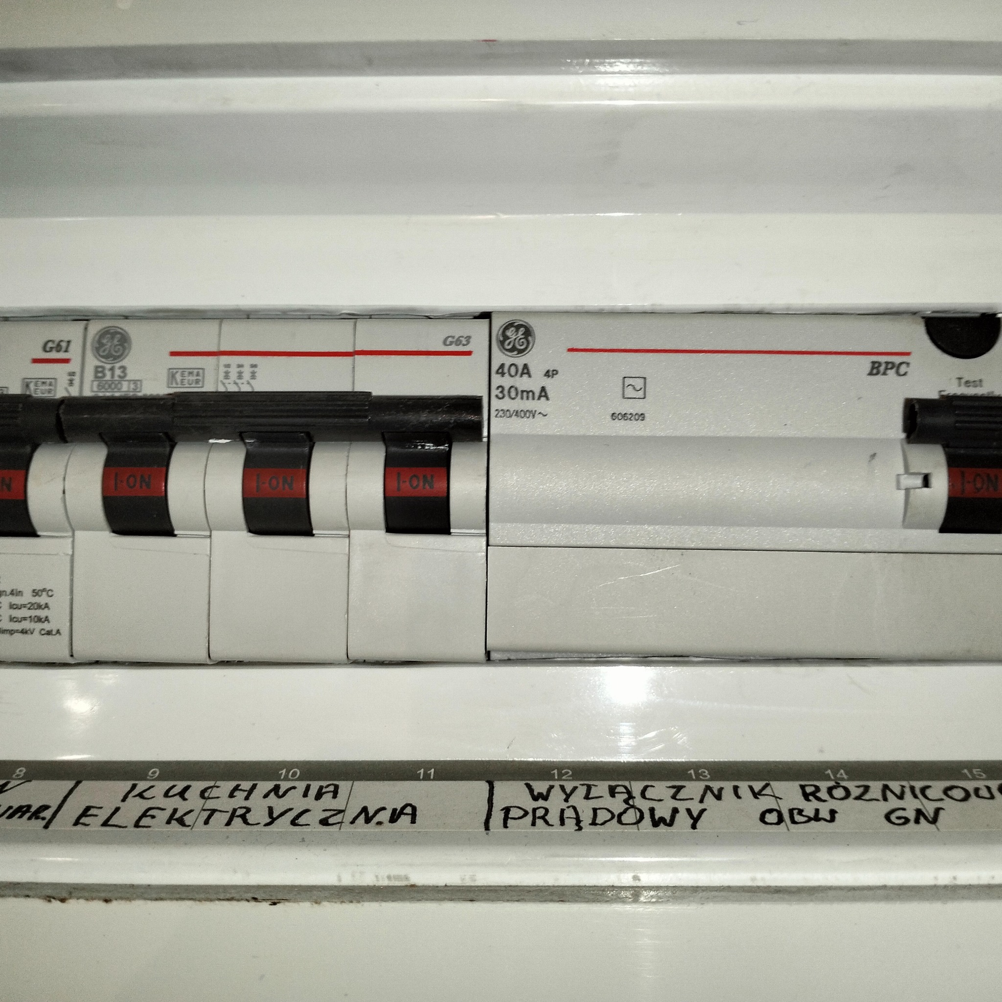 Szczegółowe ujęcie tablicy rozdzielczej z wyłącznikami, w tym wyłącznik różnicowoprądowy, z ręcznie napisanymi etykietami opisującymi obwody, takie jak 'kuchnia elektryczna'.