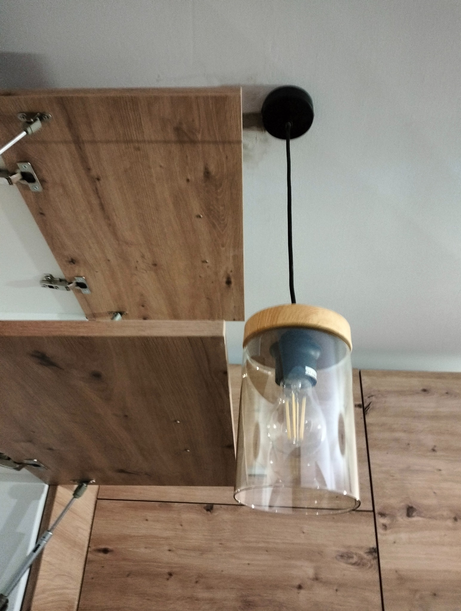 Nowoczesna lampa wisząca z elementami drewna i przezroczystym kloszem, zamontowana nad szafkami kuchennymi z drewnianymi frontami.