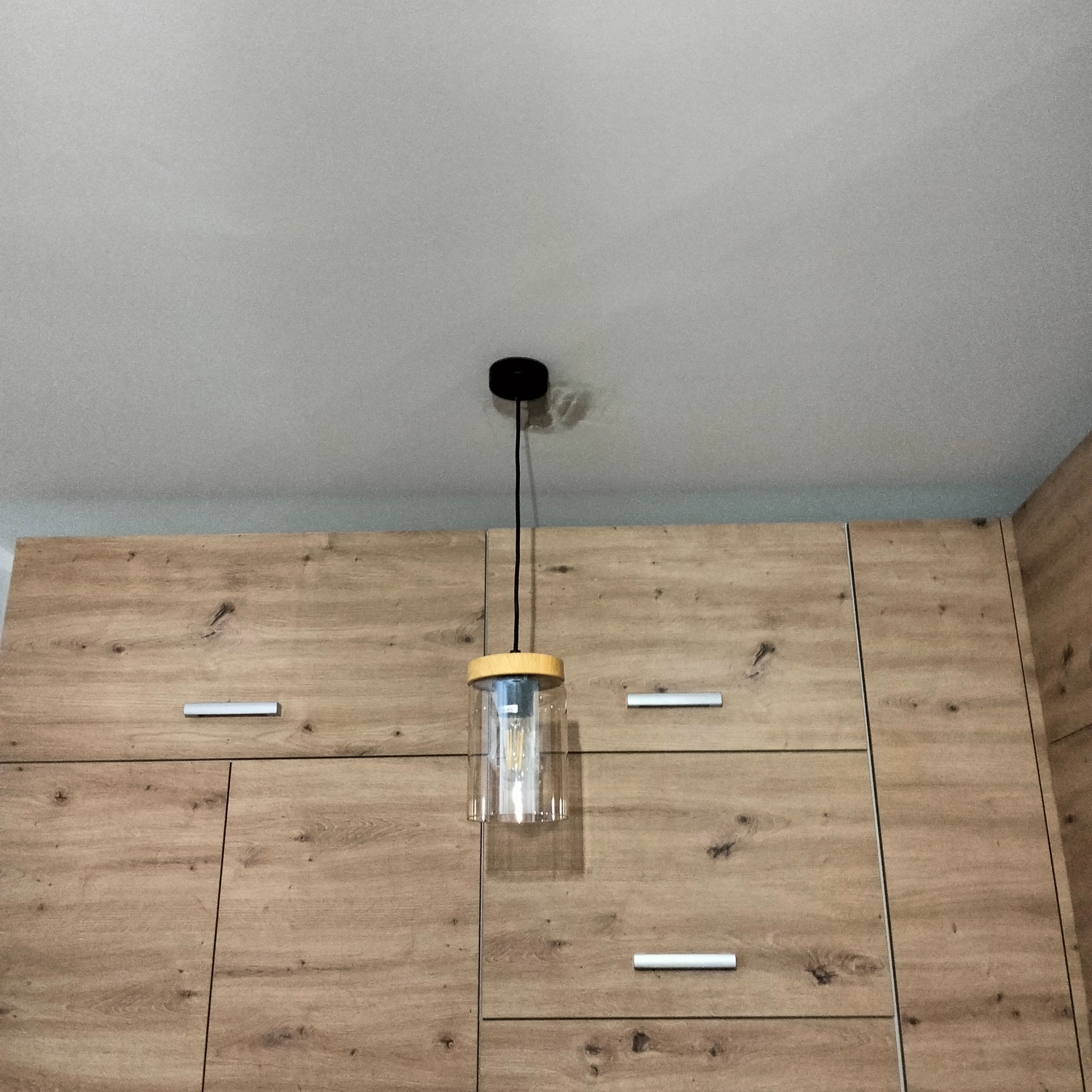 Szklana lampa wisząca z żarówką filamentową, zamontowana na tle drewnianej zabudowy meblowej.