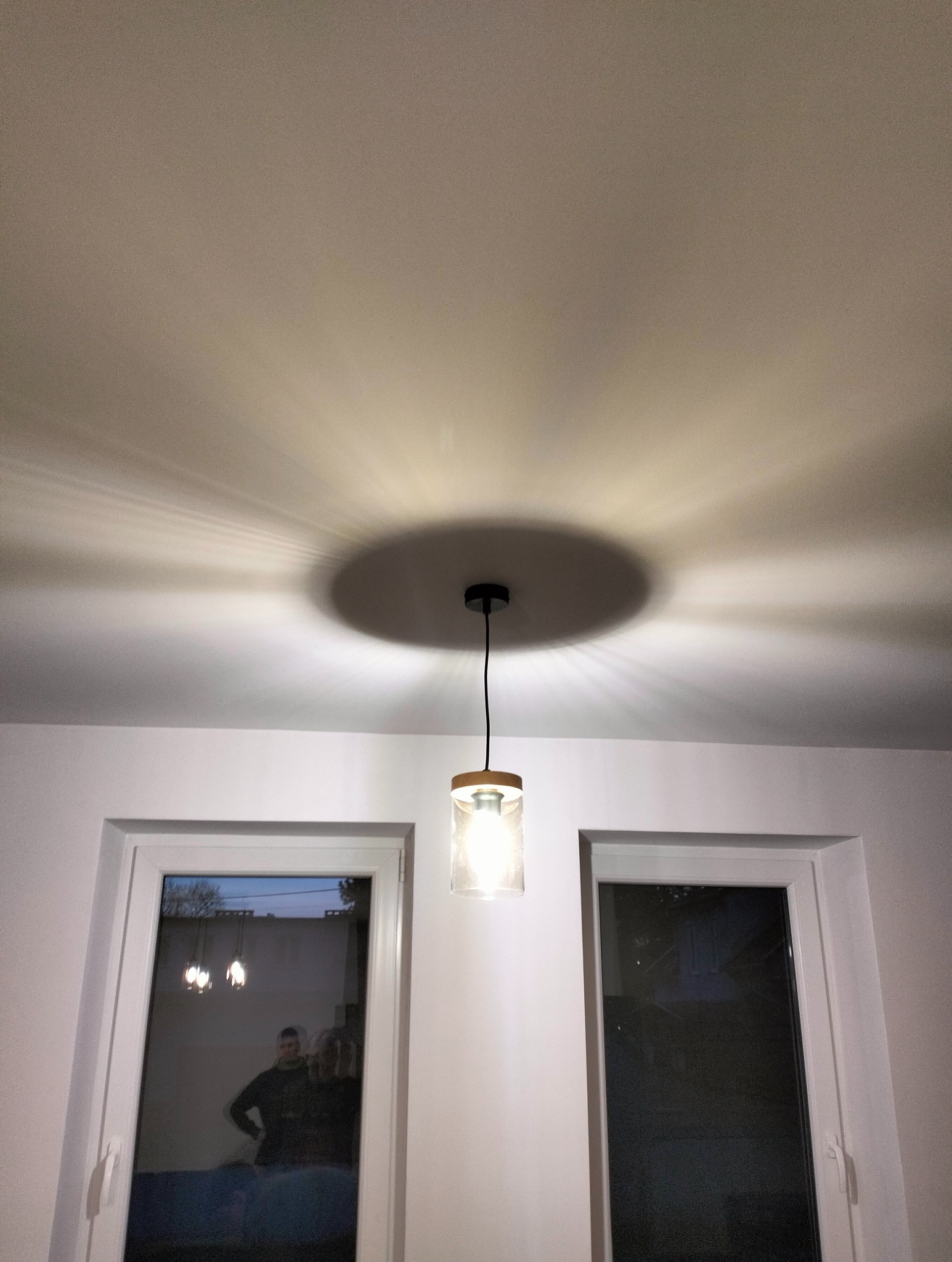 Nowoczesna lampa wisząca z cylindrycznym kloszem, oświetlająca sufit w nowym mieszkaniu, widoczna refleksja w oknie.