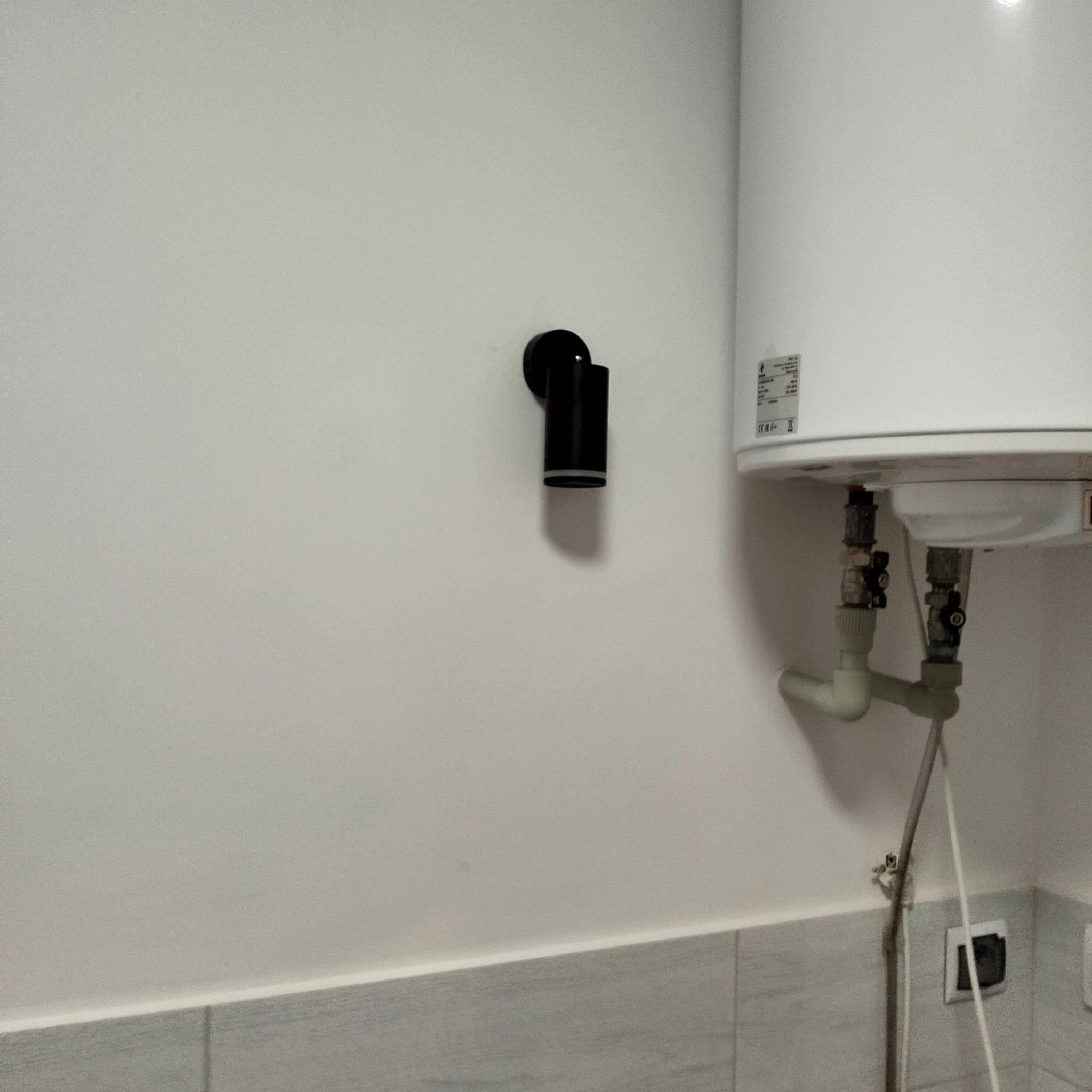 Bojler elektryczny zamontowany na ścianie w łazience, widoczne rury doprowadzające wodę i czarny minimalistyczny kinkiet LED obok.