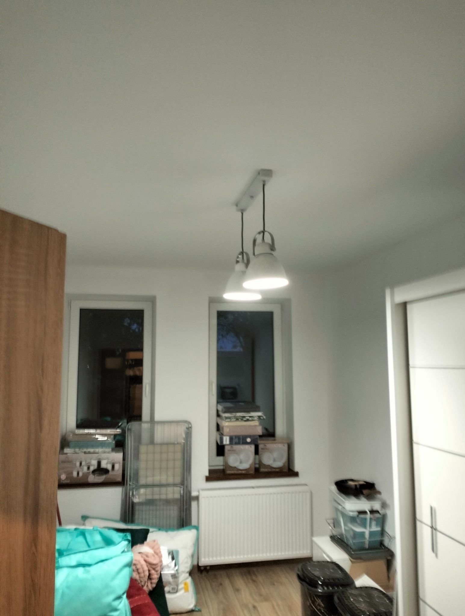 Trzy lampy wiszące z białymi kloszami w pokoju z dwoma oknami i elementami umeblowania, widok od dołu.