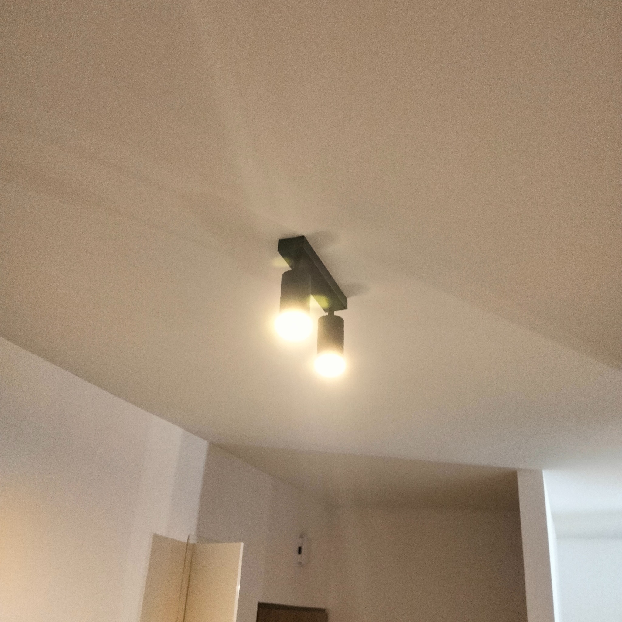 Czarna lampa sufitowa z dwoma reflektorami, świecąca ciepłym światłem na tle białego sufitu i ściany w pomieszczeniu.