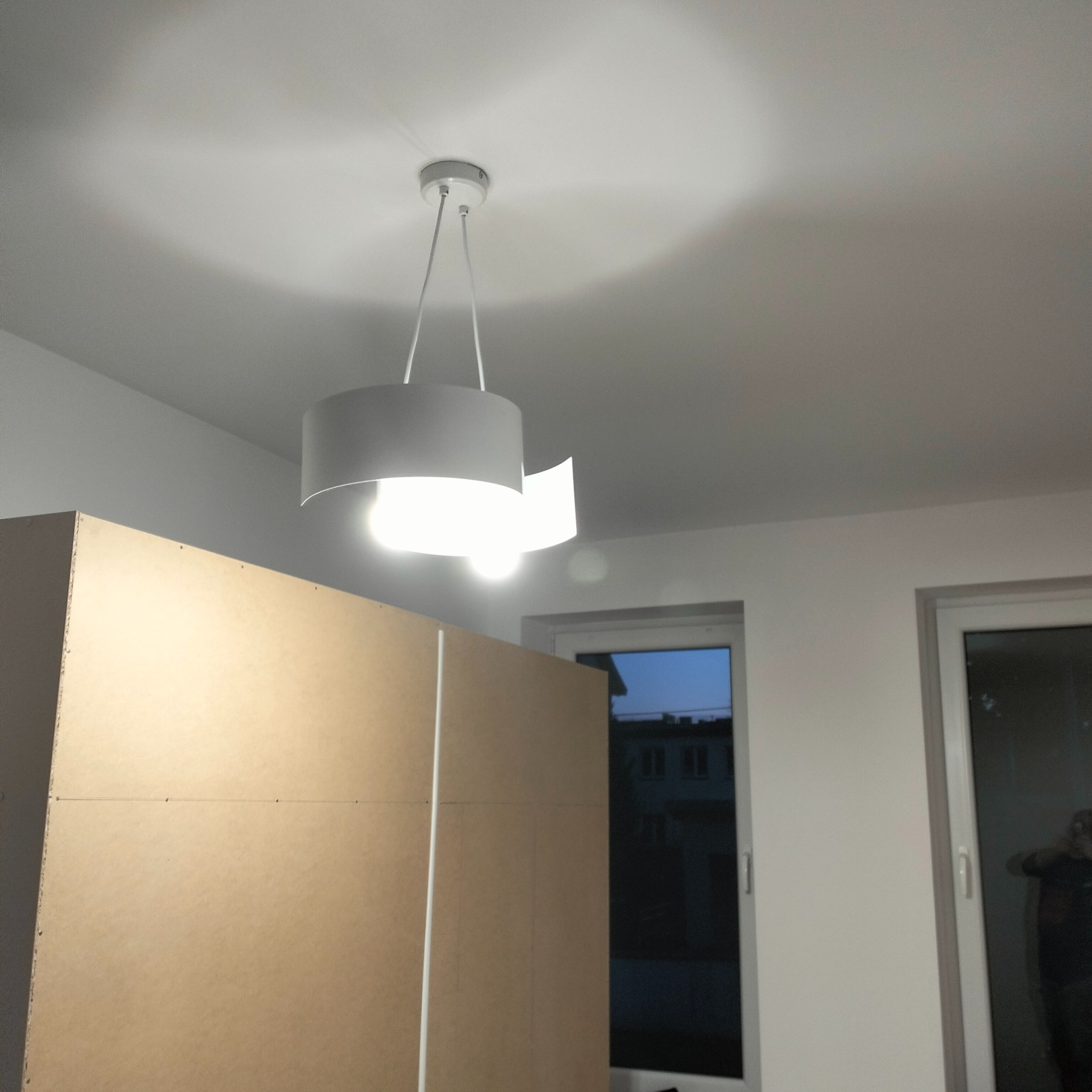 Szara lampa wisząca z dwoma kloszami, zamontowana na suficie w pomieszczeniu z oknami i ścianą z płyt kartonowo-gipsowych.