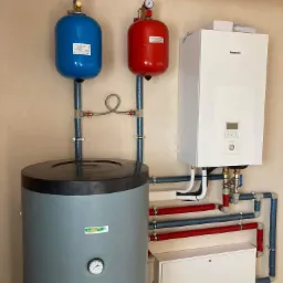 Instalacja grzewcza z zbiornikiem akumulacyjnym, dwoma naczyniami wzbiorczymi (niebieskim i czerwonym) z manometrami oraz wiszącym kotłem gazowym Panasonic, widoczne rury w izolacji termicznej.