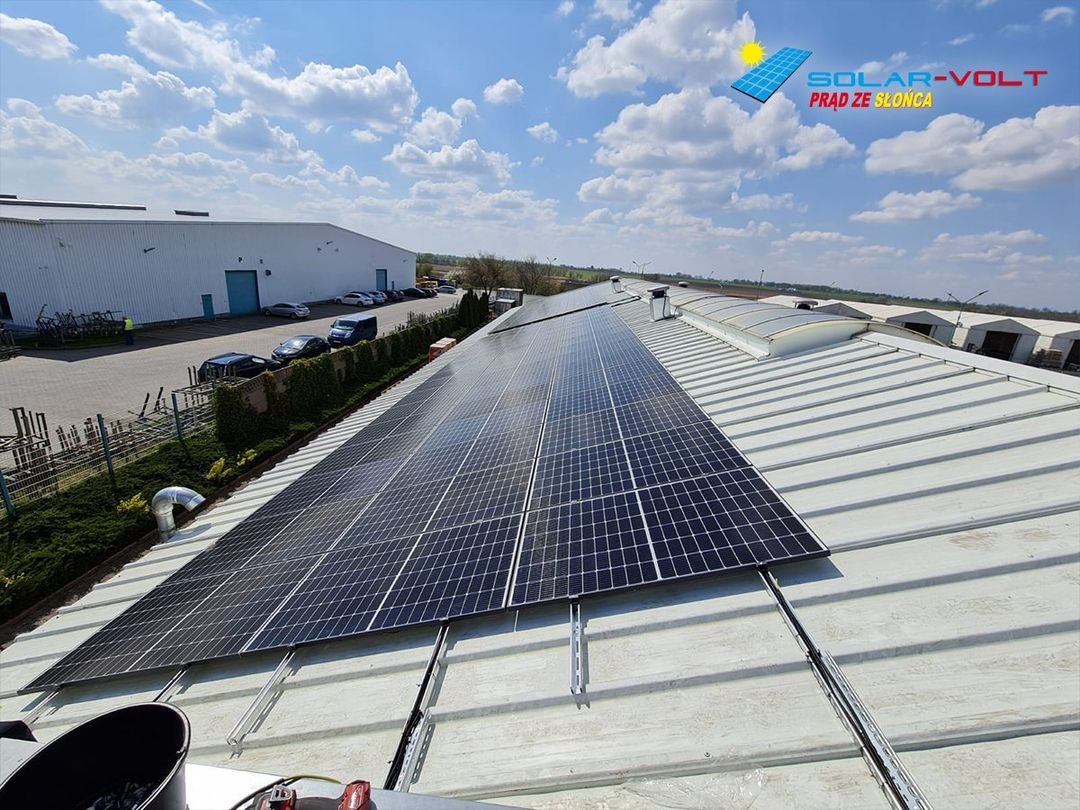 Instalacja paneli słonecznych na dachu przemysłowym z widocznymi elementami montażowymi, budynkiem w tle i błękitnym niebem z chmurami.