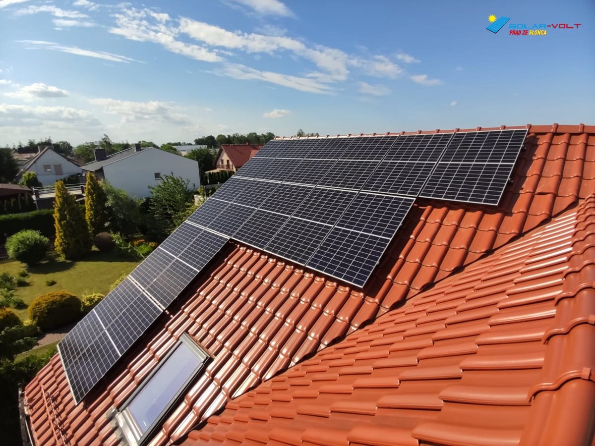 Panele słoneczne zamontowane na dachu z czerwonej dachówki ceramicznej, widoczne okno dachowe i logo firmy w prawym górnym rogu.